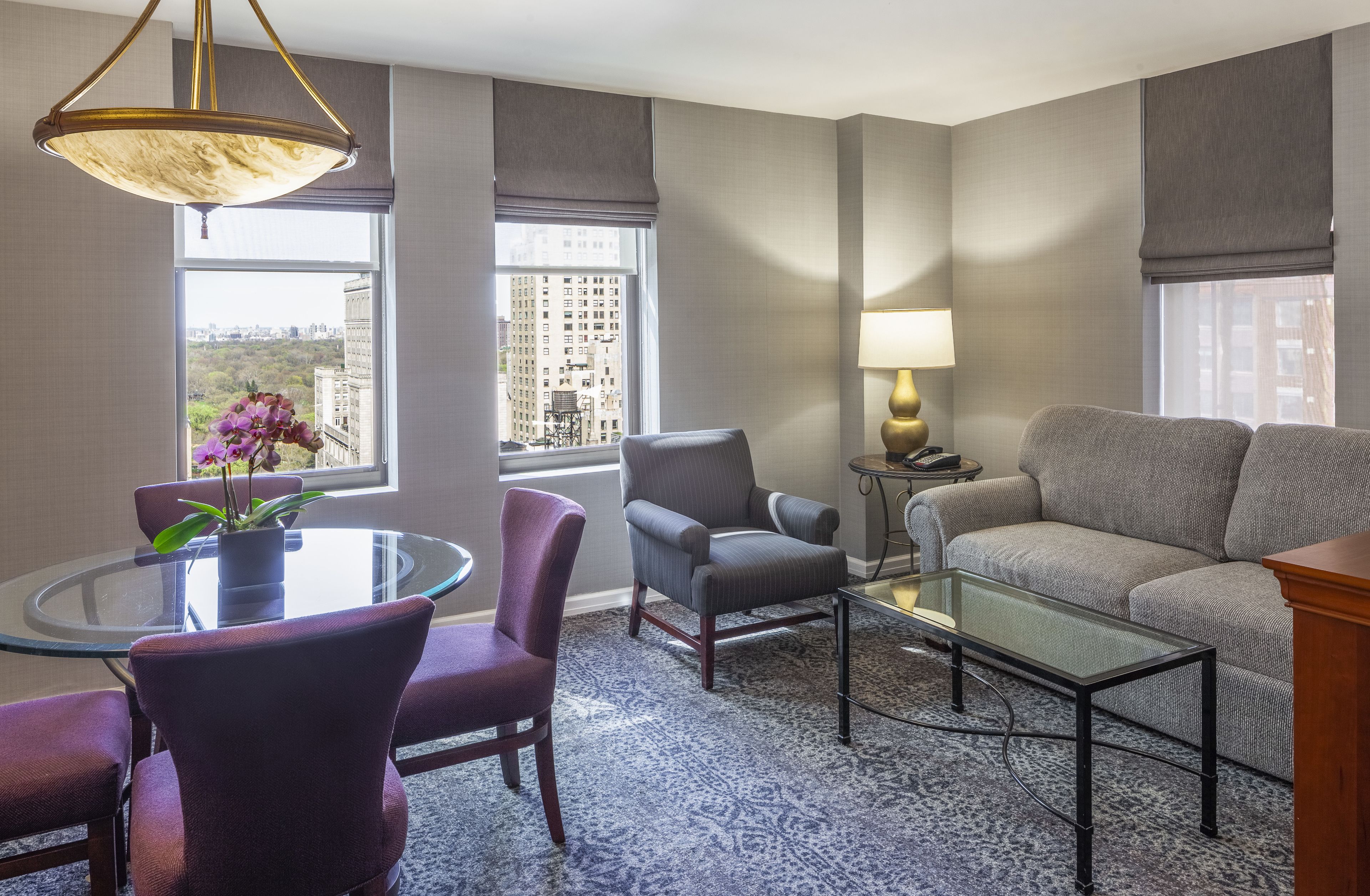 Moderni hotellihuone, jossa on pyöreä lasipöytä, violetit tuolit, harmaa nojatuoli, harmaa sohva, pieni sivupöytä lampun kanssa sekä suuret ikkunat, joista avautuu näkymä kaupunkiin.