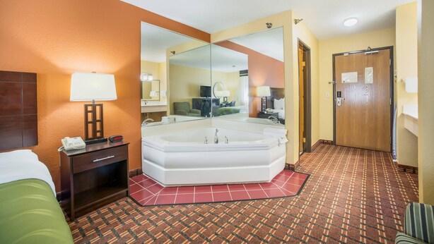 A hotel room with a large mirror, a bathtub, a bedside table with a lamp, and a green bed. - Quality Inn O'Fallon - St. Louis - Shiloh (O'Fallon)