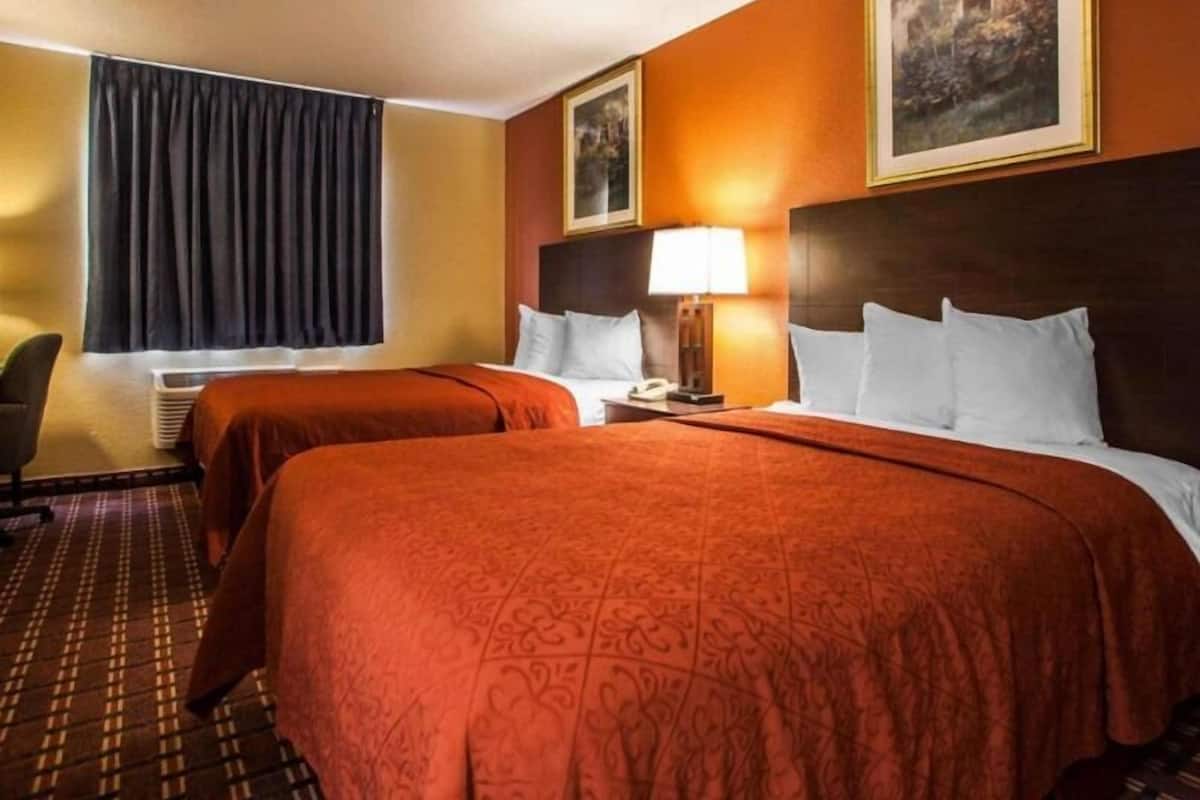 A hotel room with two beds, a desk, a chair, a lamp, and a painting on the wall.