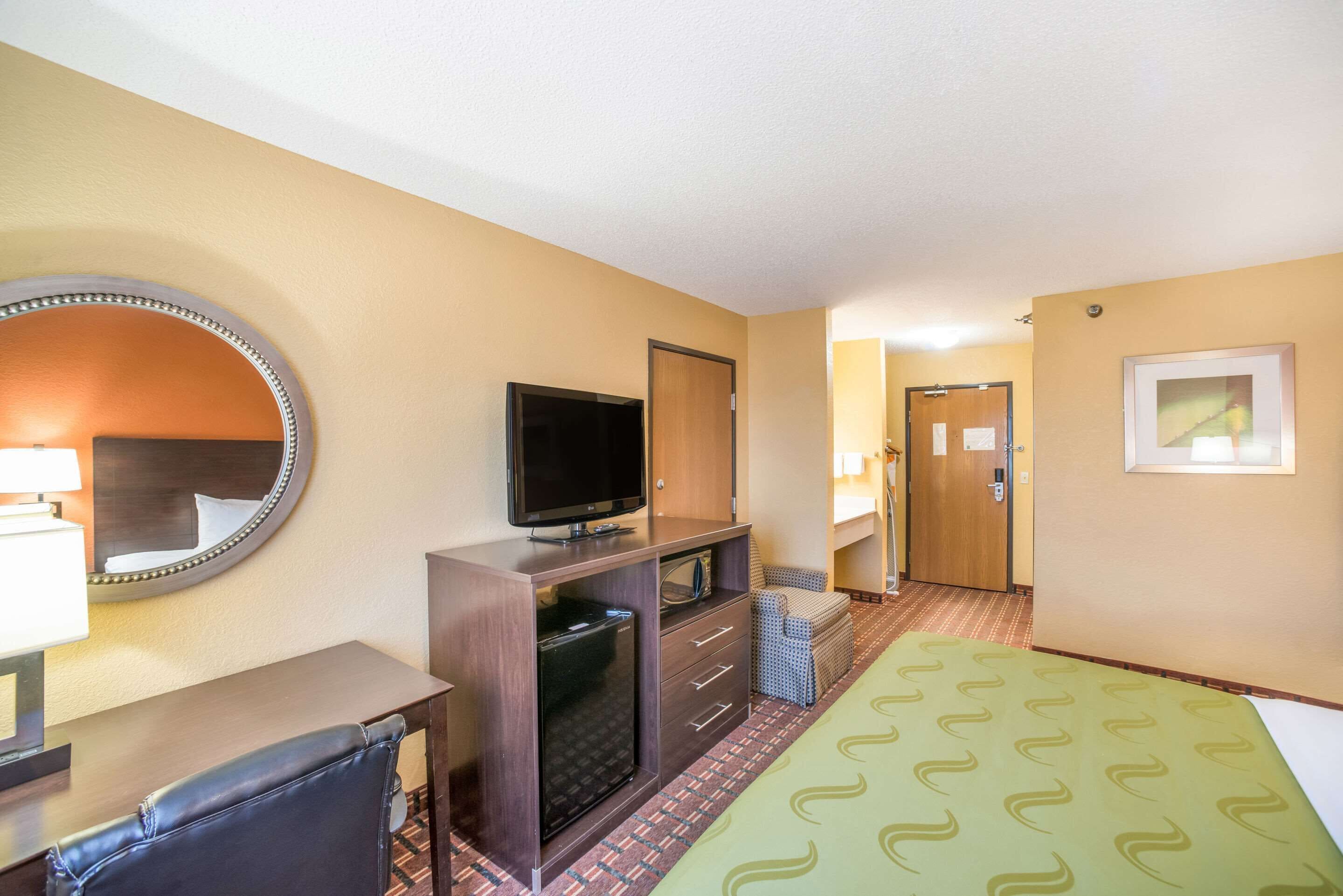 Quarto de hotel com cama, escrivaninha com televisão, espelho, cómoda e porta que dá para outro espaço.