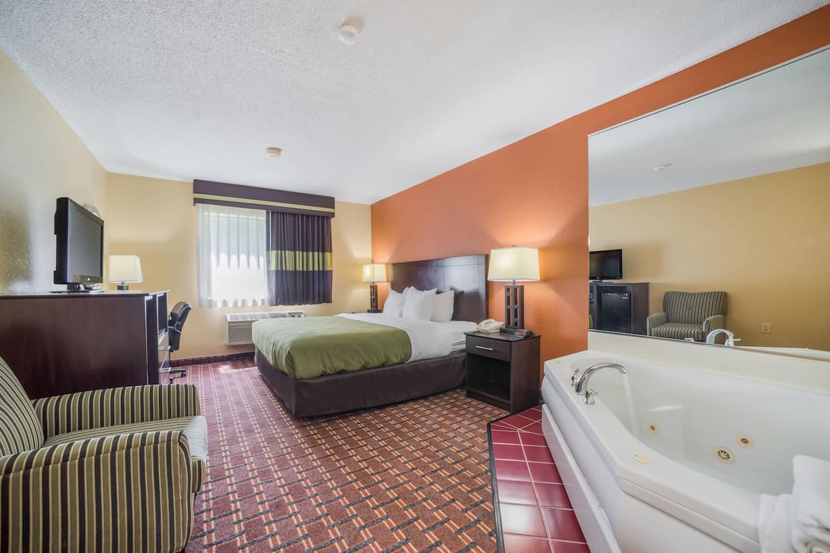 A hotel room with a bed, a desk with a computer, a chair, a large mirror, and a bathtub.