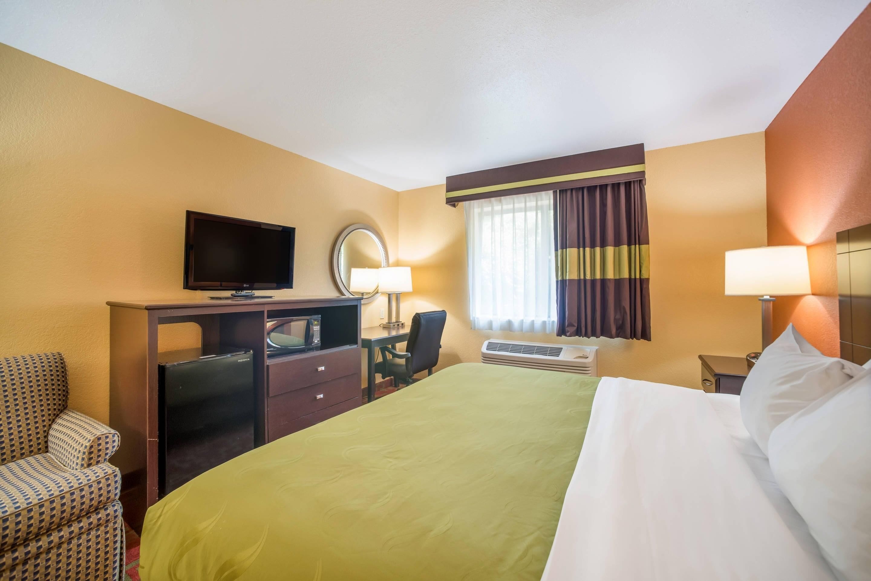 A hotel room with a bed, a desk with a computer, a television, a chair, a mirror, a window with curtains, and an air conditioning unit.