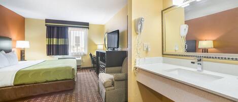 Habitación de hotel con cama, escritorio, silla, televisor, espejo y baño con lavamanos y espejo.