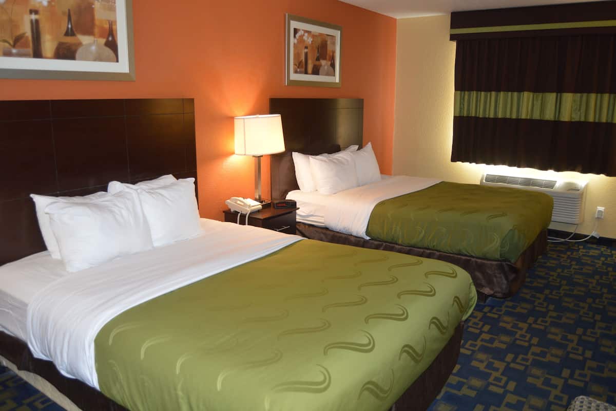 A hotel room with two beds, a nightstand with a lamp, an air conditioning unit, and artwork on the walls.