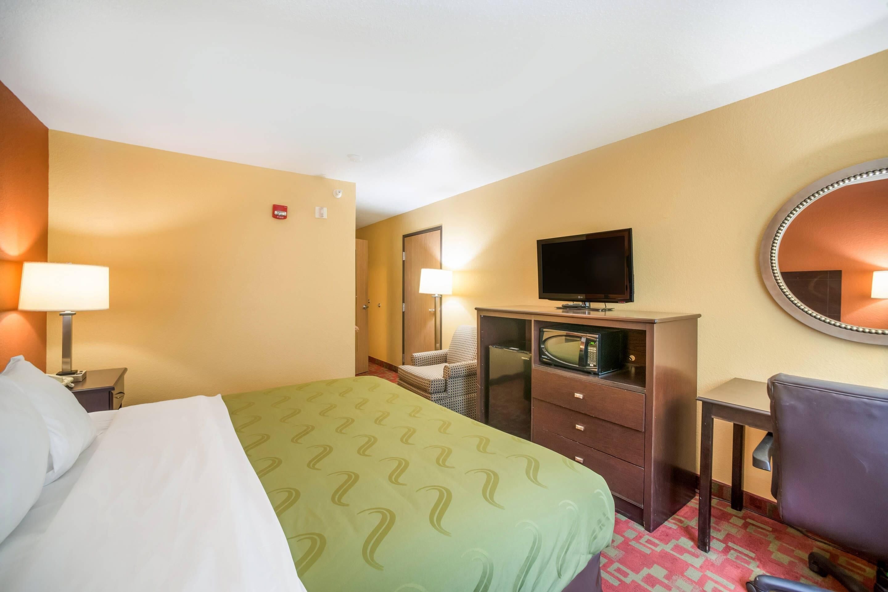 Habitación de hotel con cama, escritorio, silla, televisión, cómoda y espejo.