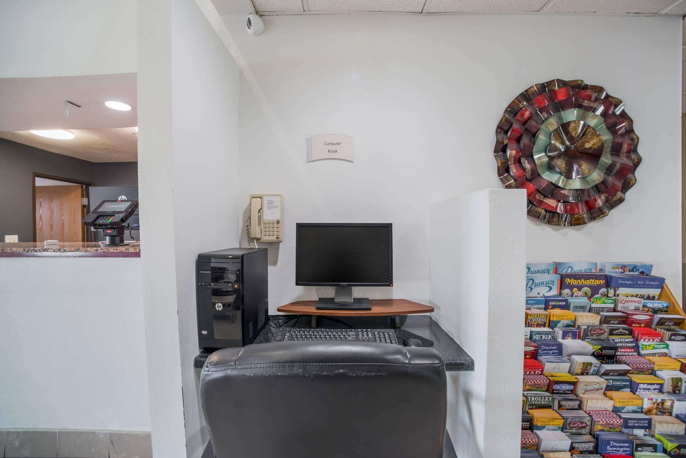 A computer desk with a monitor, keyboard, and a chair. There is a sign above the desk that reads "Computer Kiosk." A decorative metal piece is on the wall to the right.