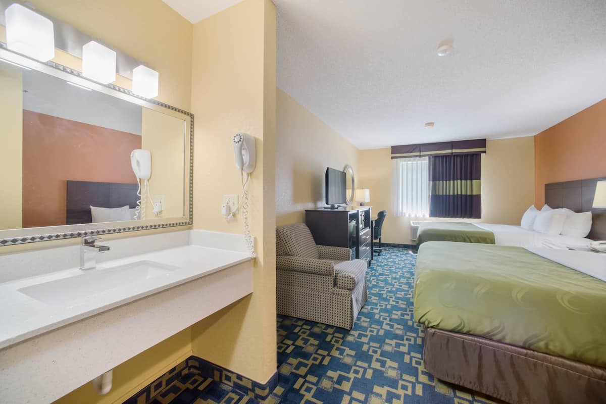 A hotel room with a large mirror, a sink, a chair, a desk, a television, and two beds.