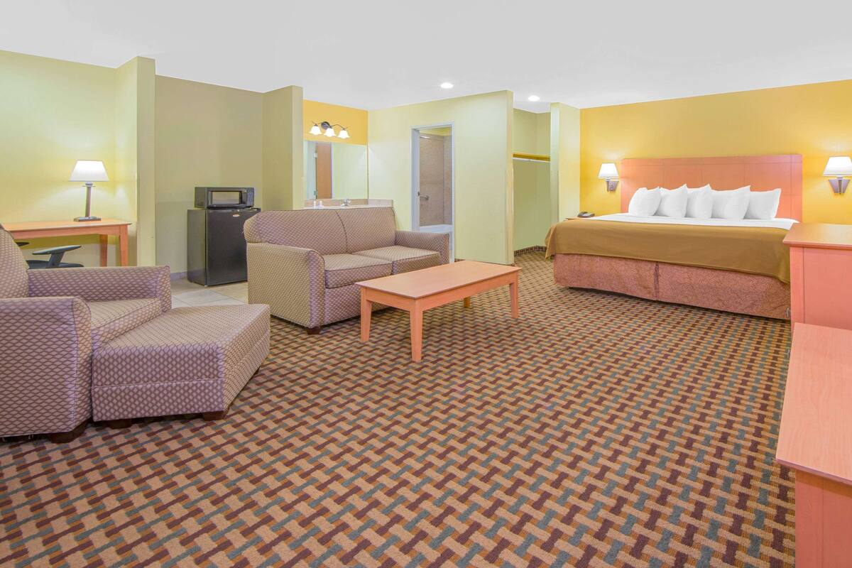 A hotel room with a bed, two armchairs, a small table, a desk with a lamp, a microwave, and a shower area.