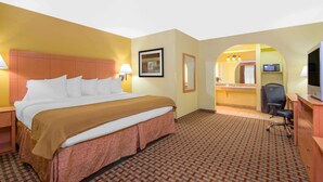 Habitación de hotel con una cama grande, un escritorio, una silla, un televisor y un baño visible a través de un arco.