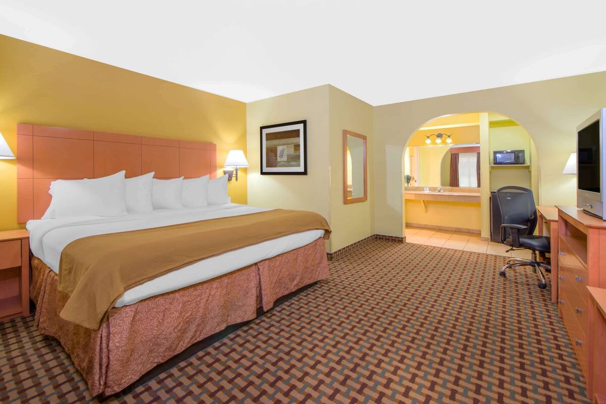 A hotel room with a large bed, a desk, a chair, a television, and a bathroom visible through an archway.