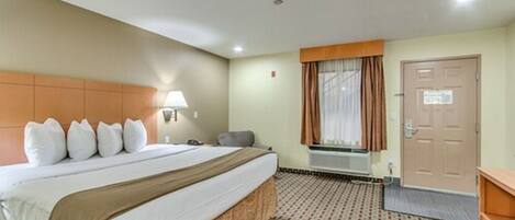 A hotel room with a bed, a desk, a chair, a window with curtains, a door, and a patterned carpet.
