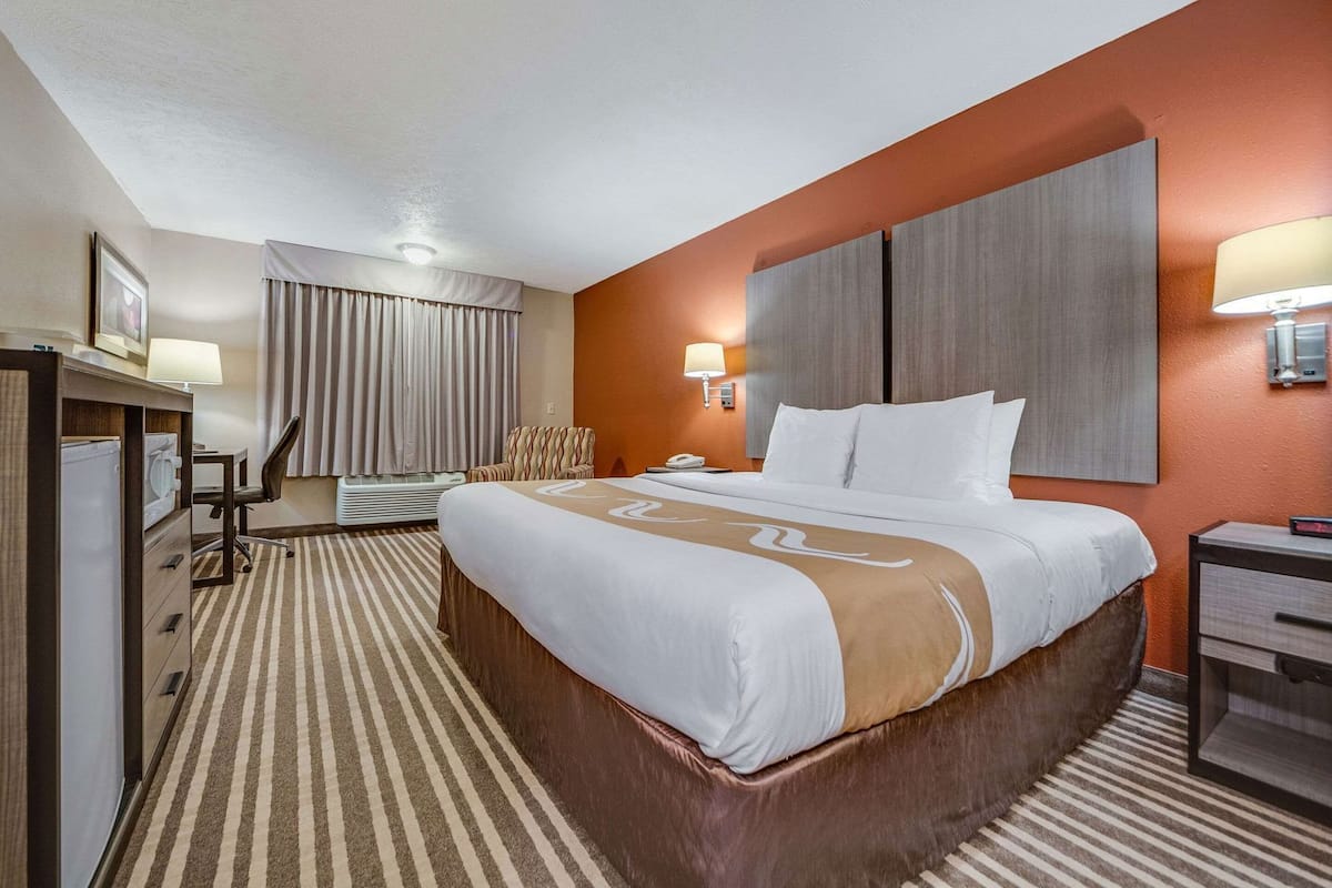 A hotel room with a large bed, a desk with a chair, a desk lamp, a telephone, and a wall-mounted lamp.
