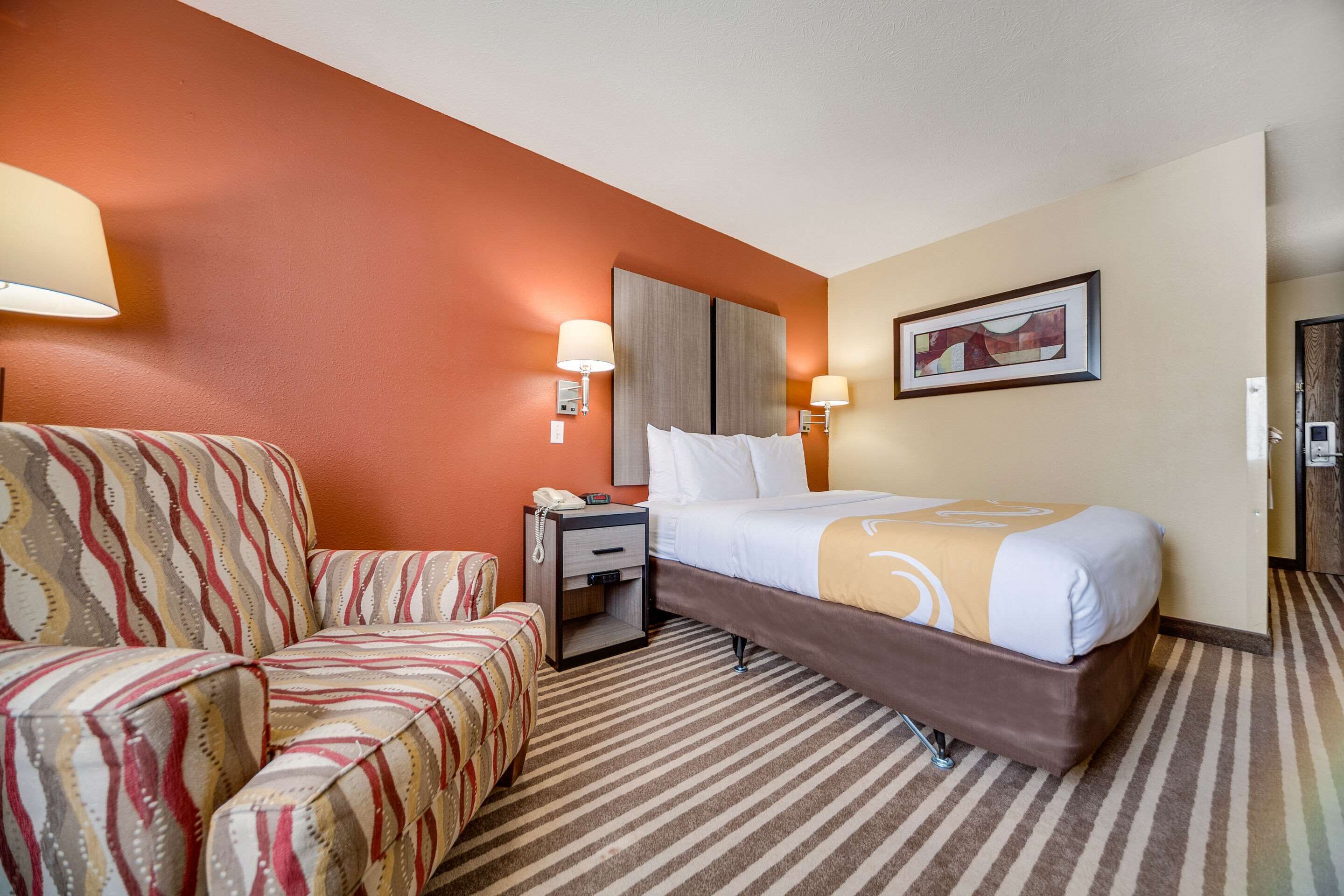 Una habitación de hotel con una cama, una mesita de noche, una silla y una lámpara de pared.
