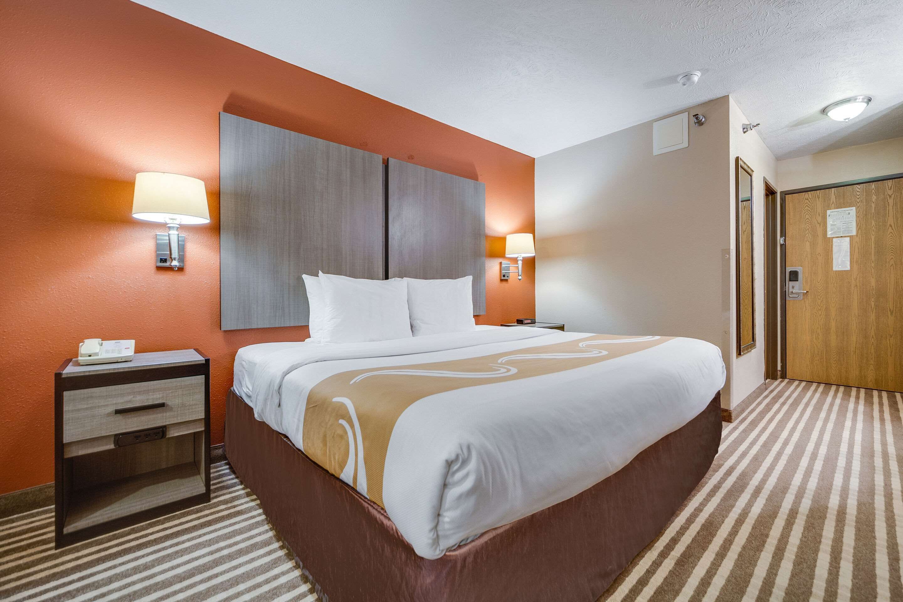 Una habitación de hotel con una cama grande, una mesita de noche, una lámpara y una puerta.