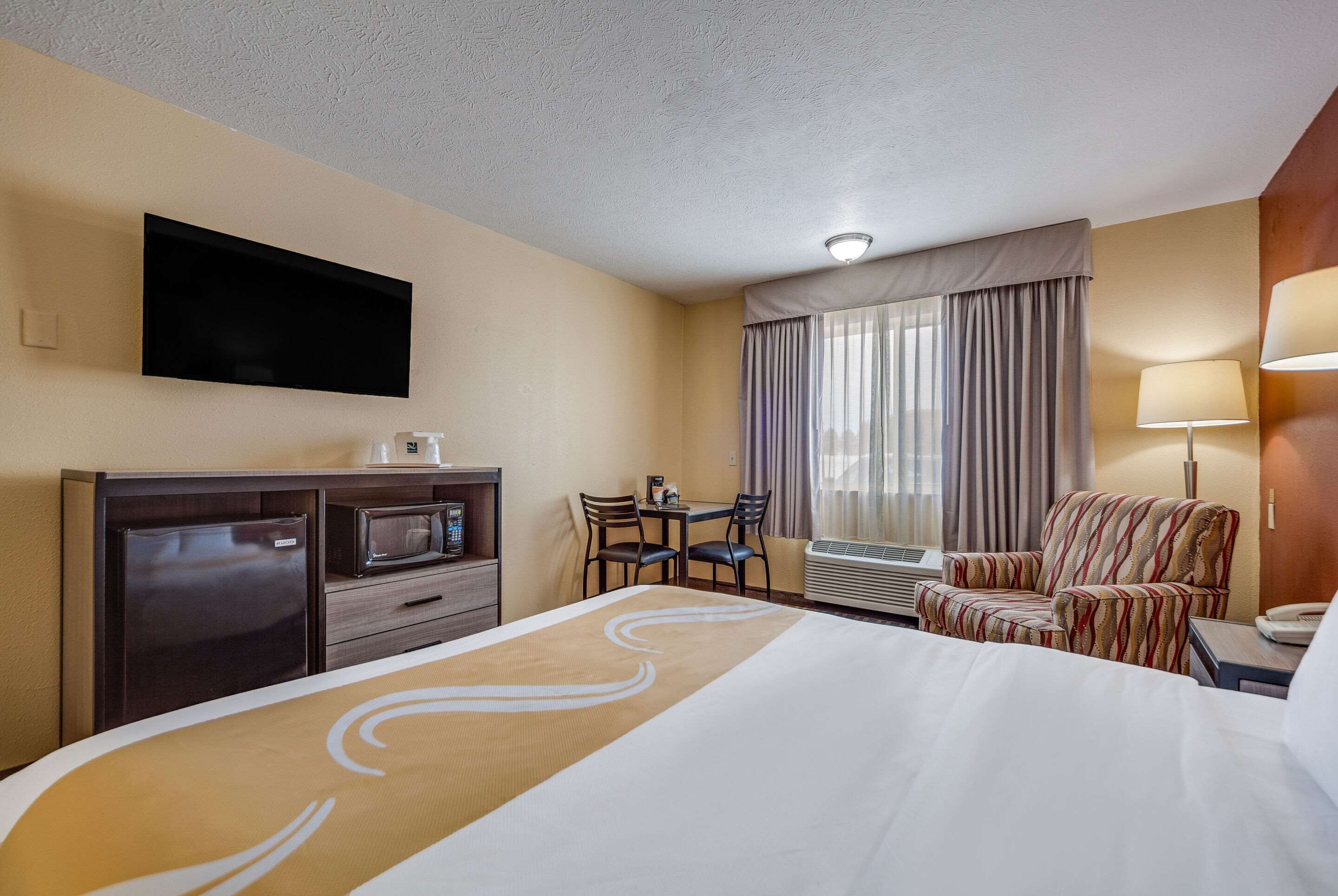 Habitación de hotel con cama, escritorio con silla, televisor montado en la pared y ventana con cortinas.