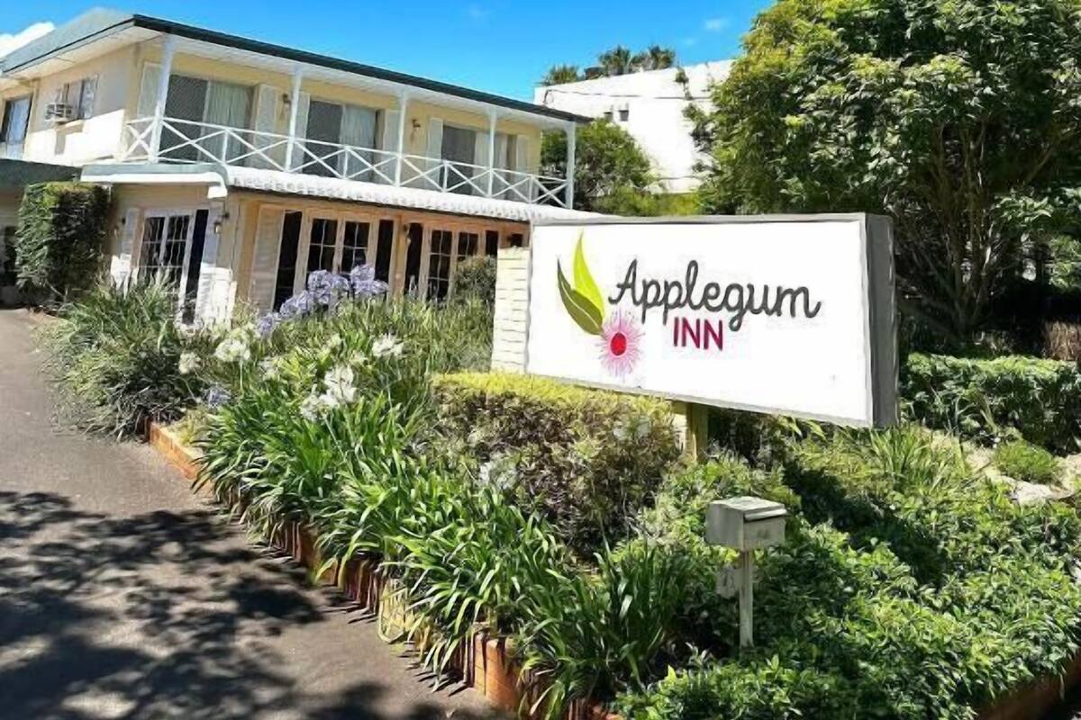 Un hotel llamado 'Applegum Inn' con un letrero, rodeado de abundante follaje y un cielo azul despejado.