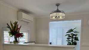 A reception area with a white counter, a chandelier, a potted plant, and a window with blinds.
