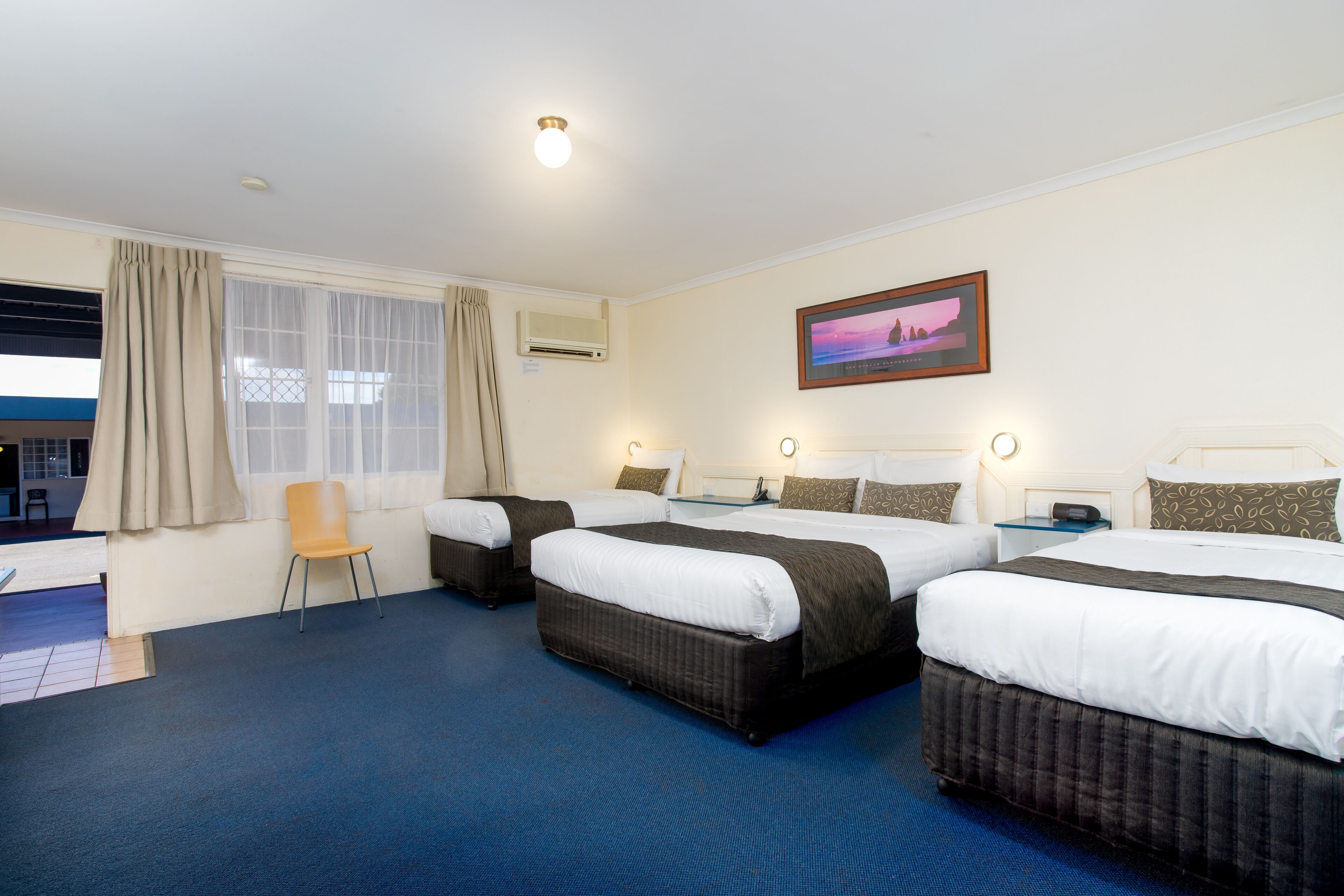 A hotel room with three beds, a chair, a painting, and an air conditioning unit.