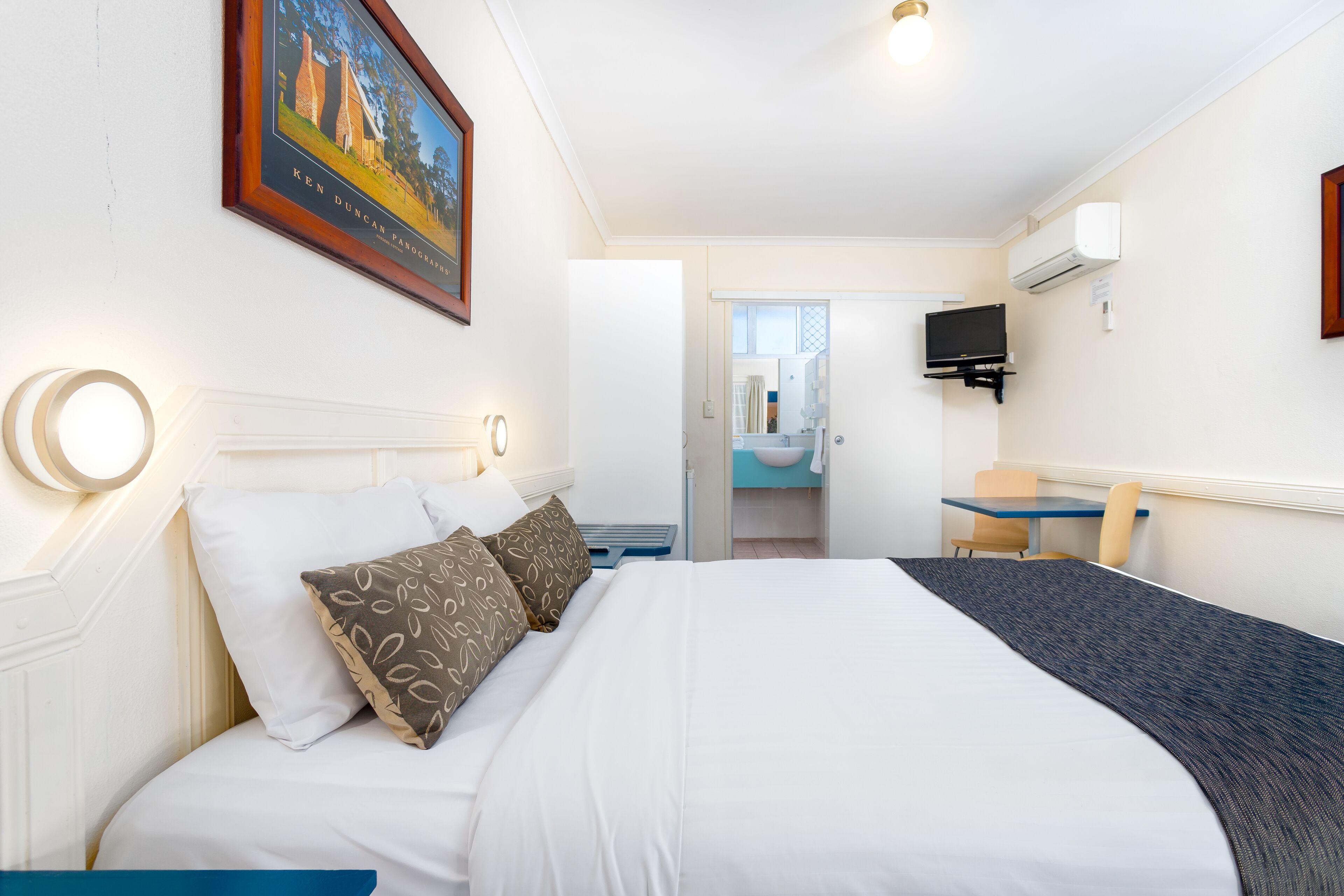 A hotel room with a bed, a desk, a chair, a television, and a bathroom visible through an open door.