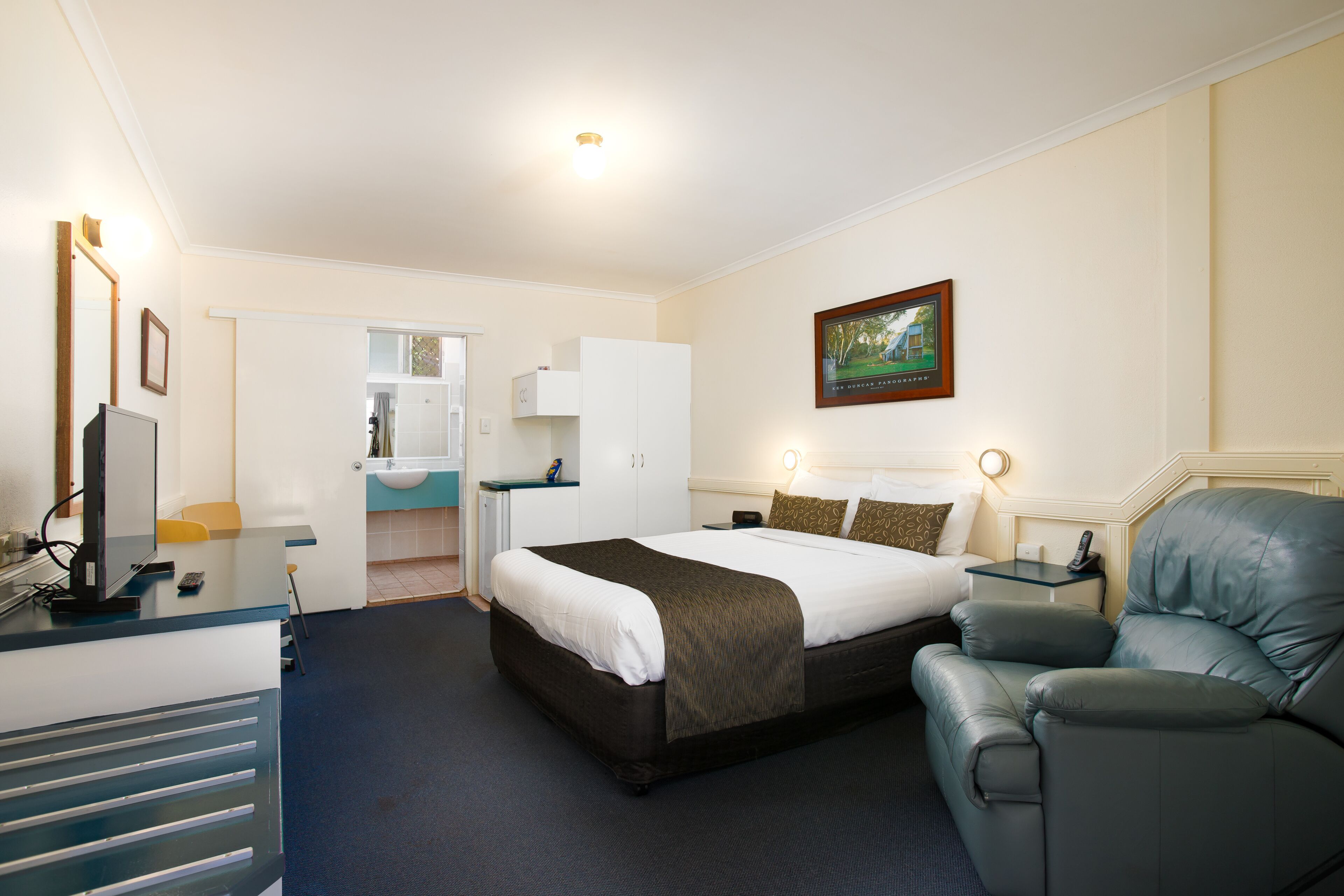 Une chambre d’hôtel avec un lit, un bureau avec un ordinateur, une chaise, une table de chevet et un tableau au mur.