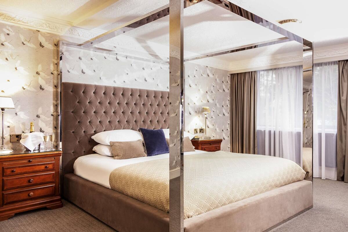 A four-poster bed with a tufted headboard, a wooden nightstand with a lamp, and a wall-mounted clock.