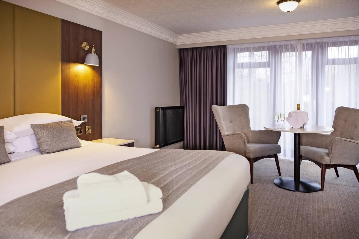 A hotel room with a large bed, two armchairs, a small table with glasses, and a radiator.