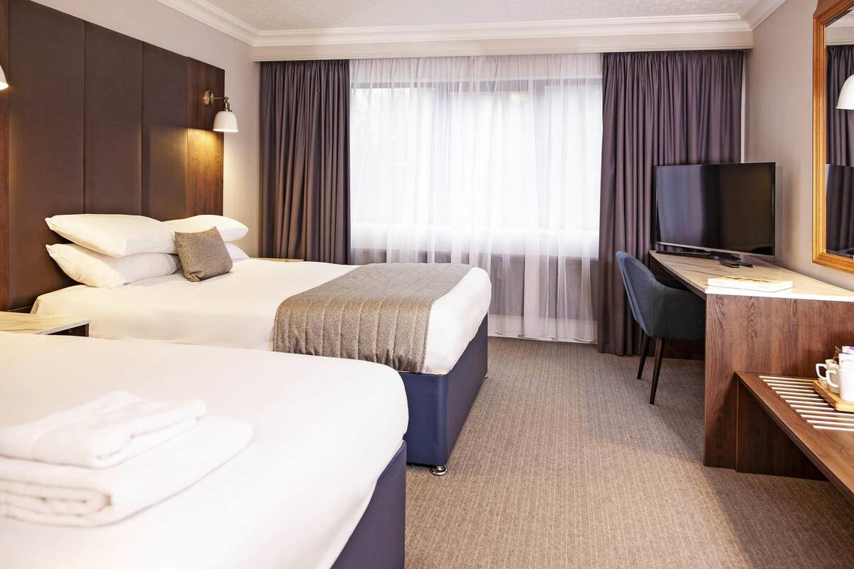 A hotel room with two beds, a desk, a television, and a large window with curtains.