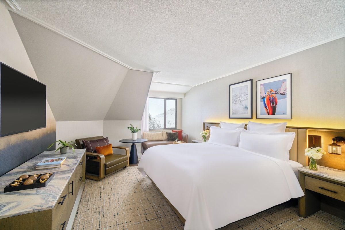 A hotel room with a large bed, a flat-screen TV, a seating area with a sofa and armchair, a small table, and artwork on the walls.