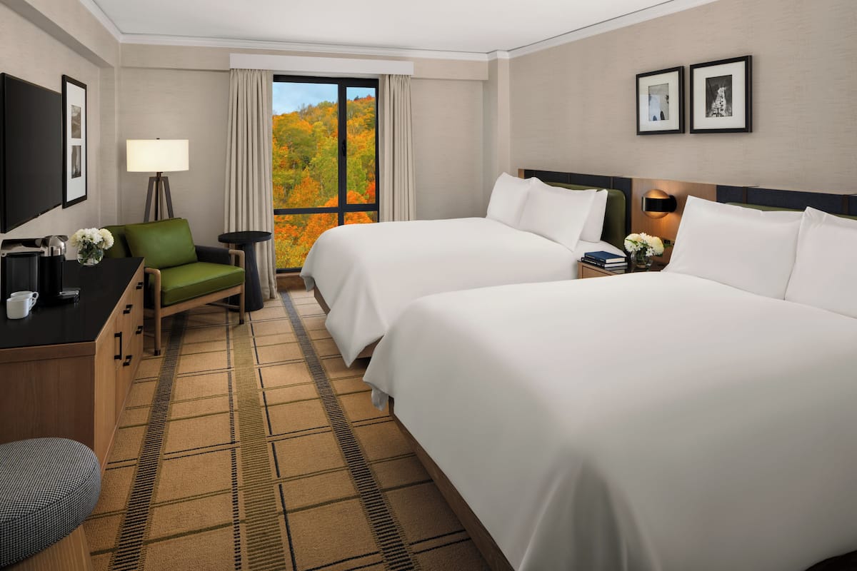A hotel room with two beds, a desk with a chair, a TV, and a window with a view of autumn foliage.