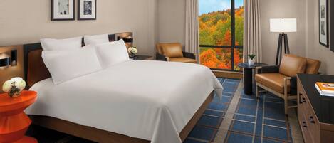 A hotel room with a large bed, a desk, a chair, a lamp, and a view of autumn foliage through the window.