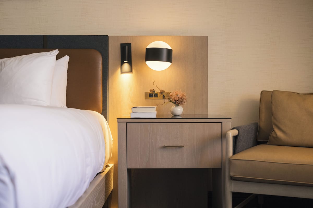A hotel room with a bed, a nightstand with a lamp, a vase with flowers, and a chair.