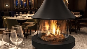 A restaurant with a central fire feature, tables with chairs, and a patterned floor. - Fairmont Tremblant (Mont-Tremblant)