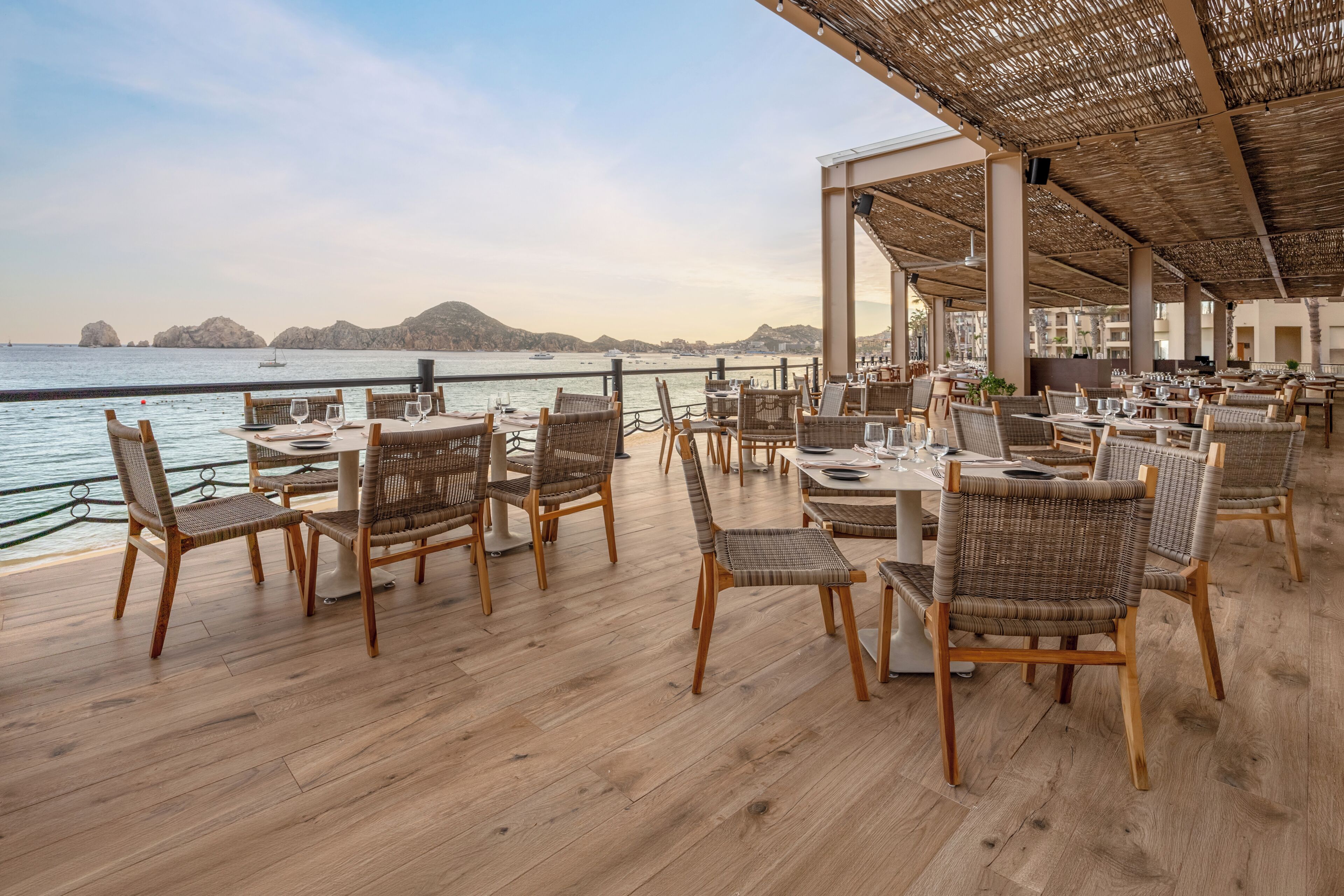 An outdoor dining area with wicker chairs and tables, overlooking a body of water with boats.