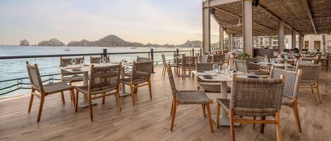 An outdoor dining area with wicker chairs and tables, overlooking a body of water with boats.