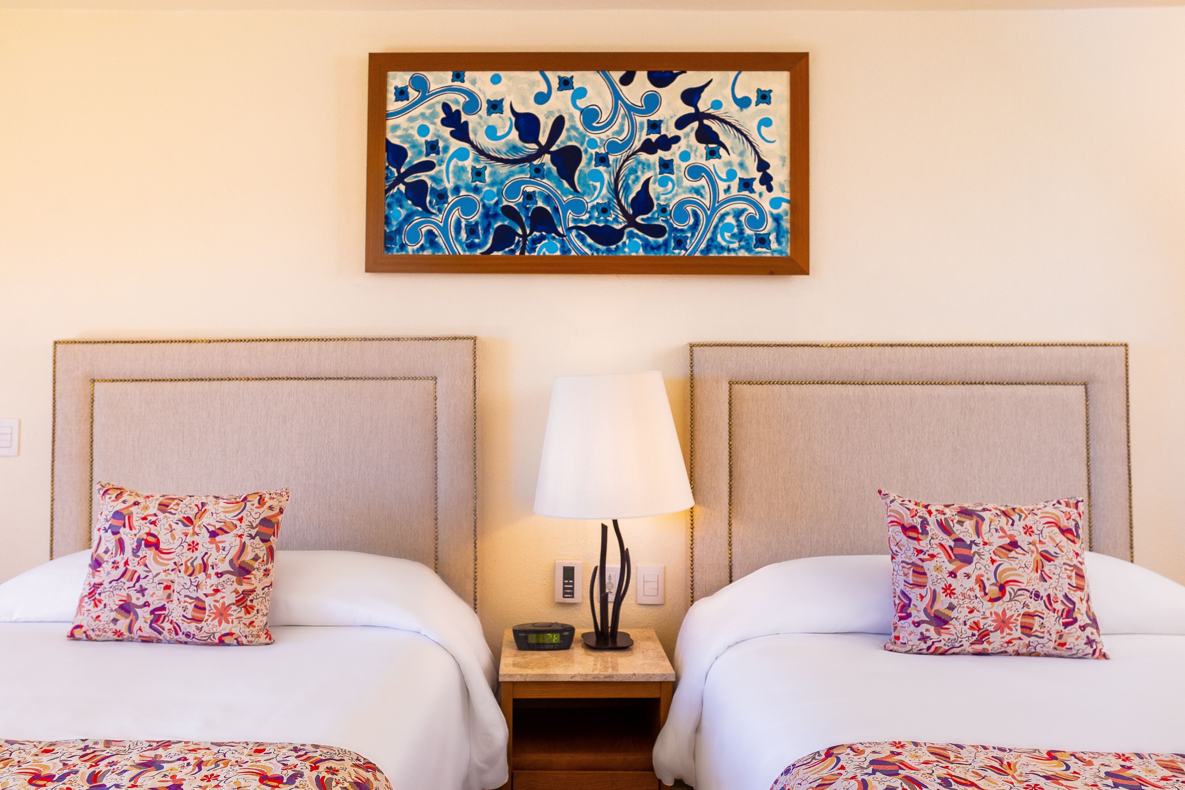 Ein Hotelzimmer mit zwei Betten, jedes mit einem gemusterten Kissen, einem Nachttisch mit Lampe und einem an der Wand hängenden Gemälde.