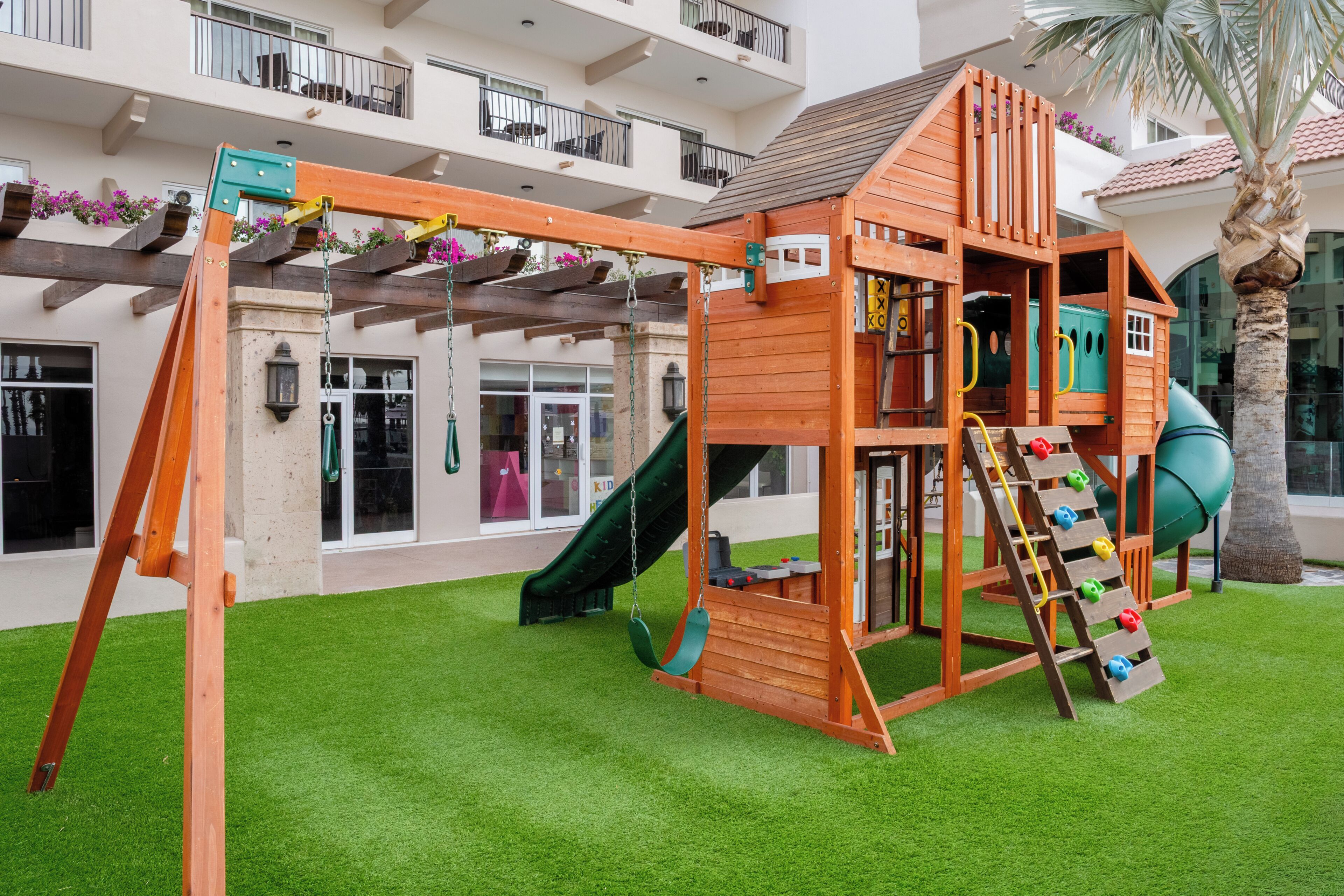 A wooden playhouse with a slide and climbing net, surrounded by artificial grass, in front of a building with balconies and windows.