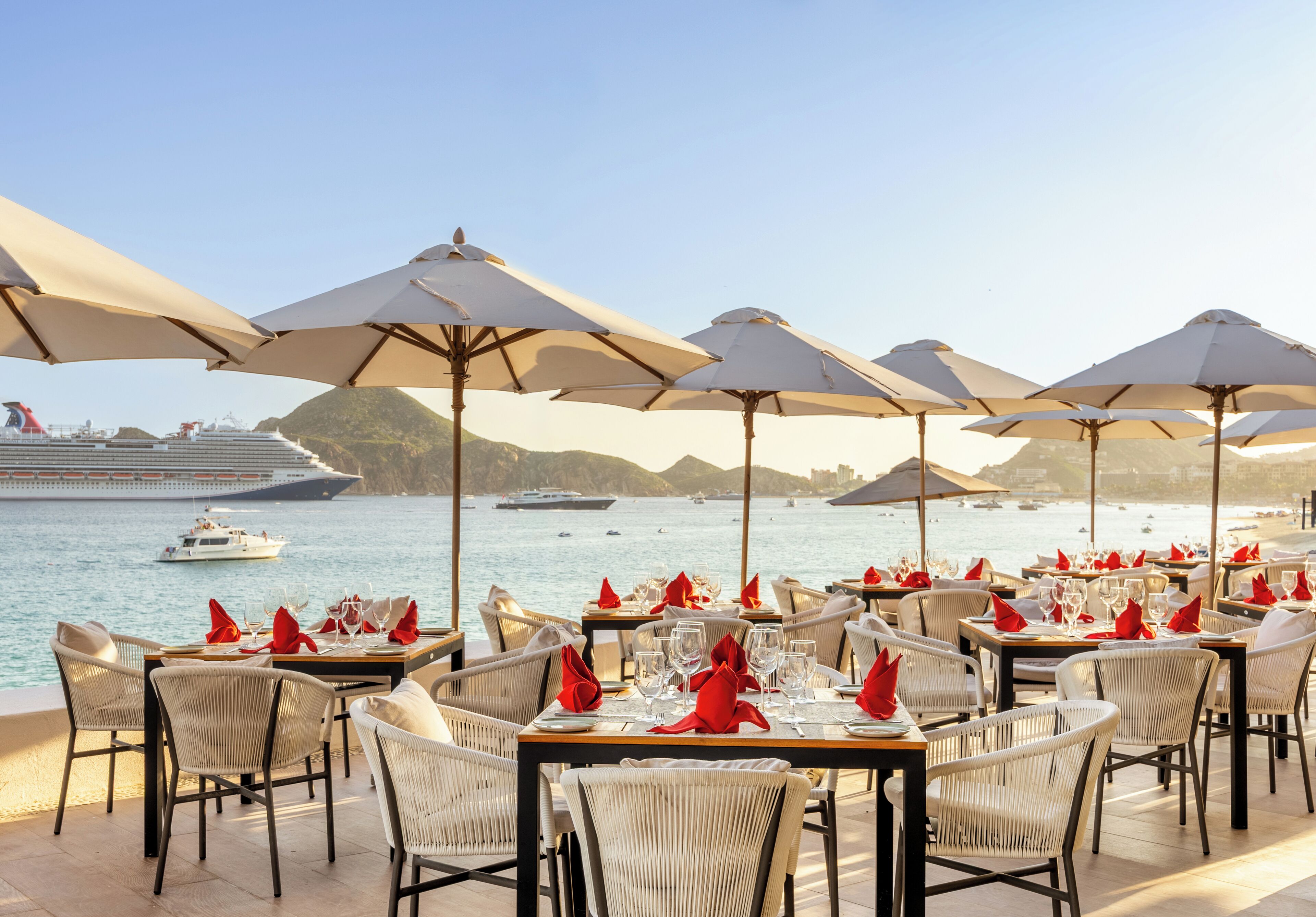 A seaside restaurant with tables set for dining, overlooking the ocean and a cruise ship.