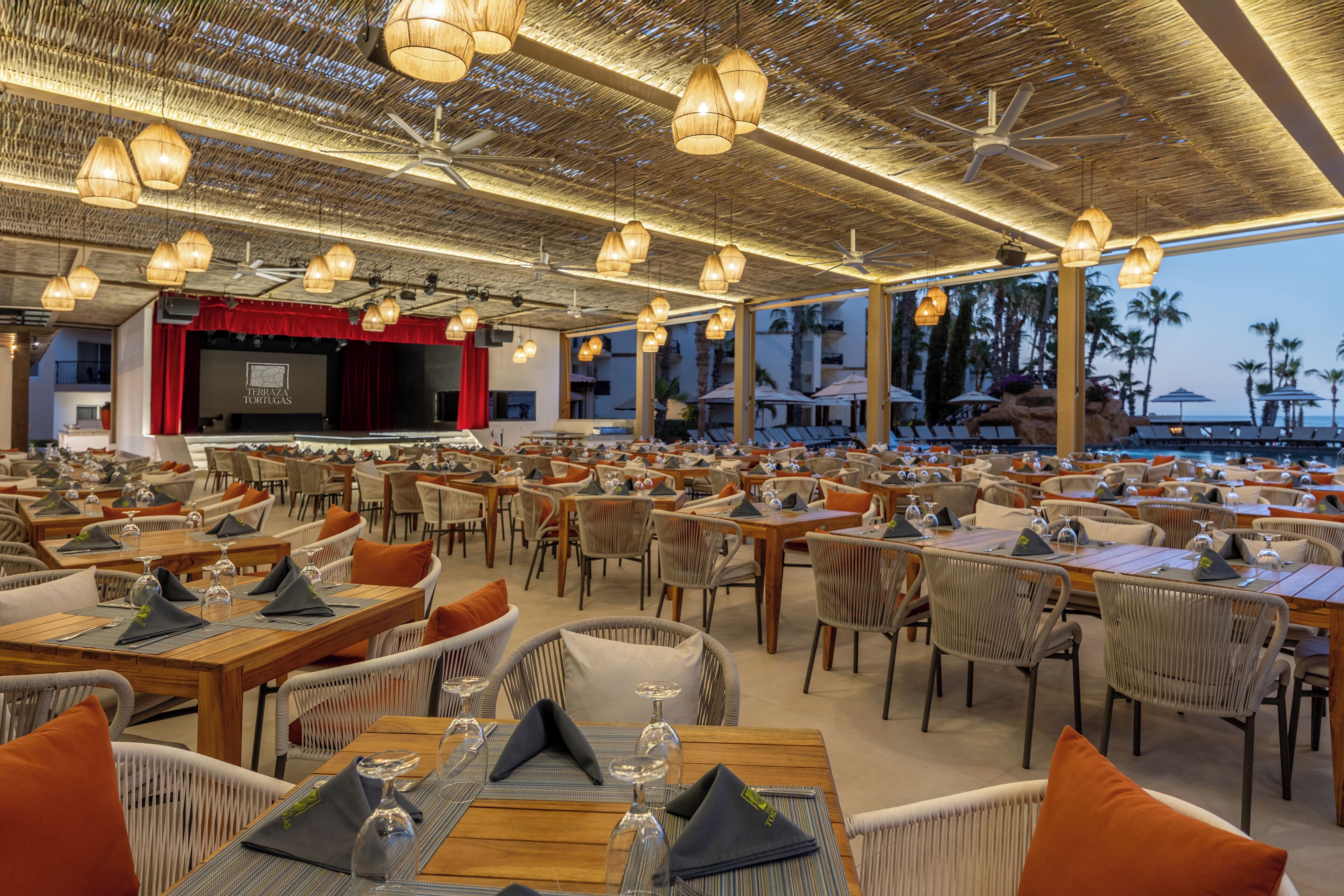 A spacious outdoor restaurant with wooden tables and wicker chairs, a stage with a red curtain, and a view of palm trees and a pool.