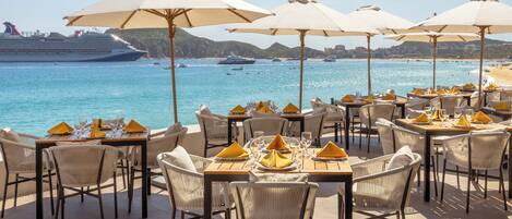 A seaside restaurant with outdoor seating, tables with umbrellas, and a view of the ocean and cruise ship.