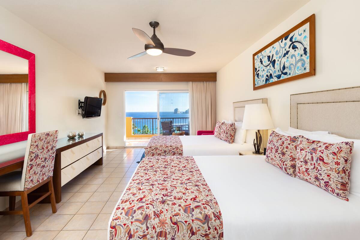 A hotel room with two beds, a desk, a chair, a ceiling fan, a TV, a balcony with a view, and a painting on the wall.