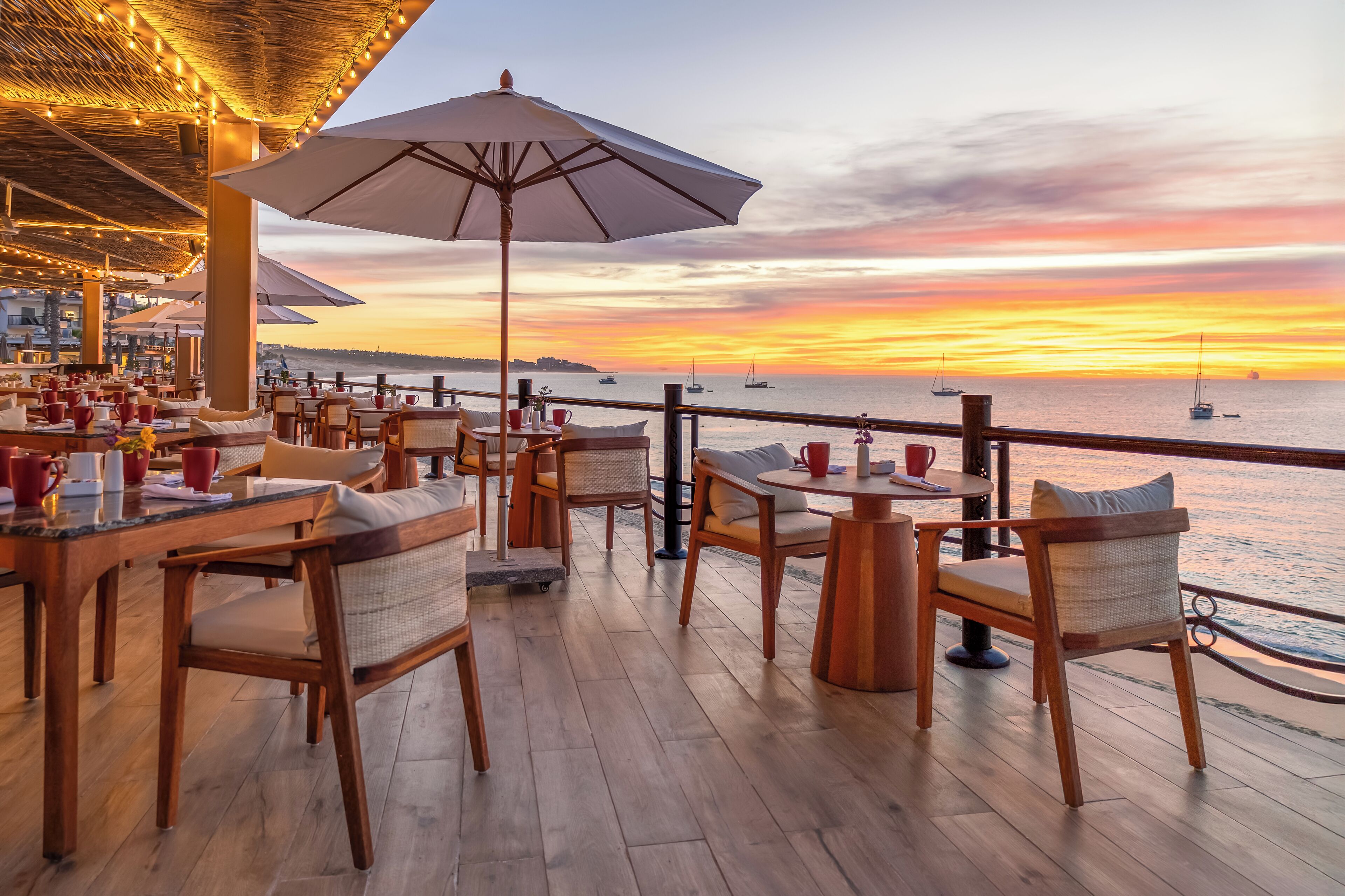 Un restaurante frente al mar con mobiliario de madera, sombrillas y vistas al océano al atardecer.