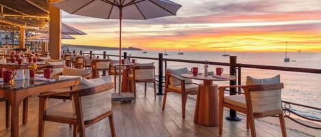 Un restaurante frente al mar con mobiliario de madera, sombrillas y vistas al océano al atardecer.