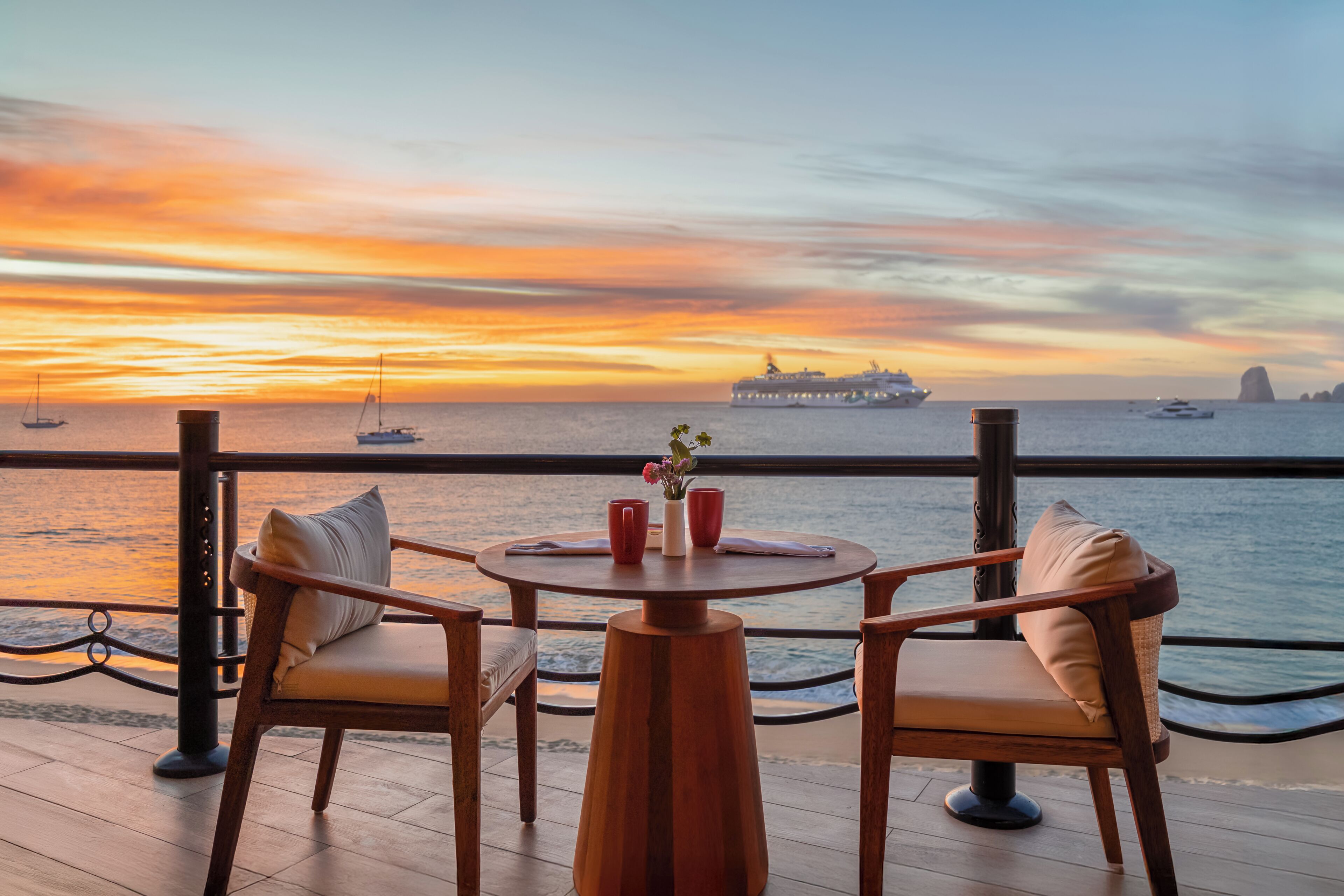 A wooden deck with two chairs and a table, overlooking a sunset over the ocean with ships in the distance.
