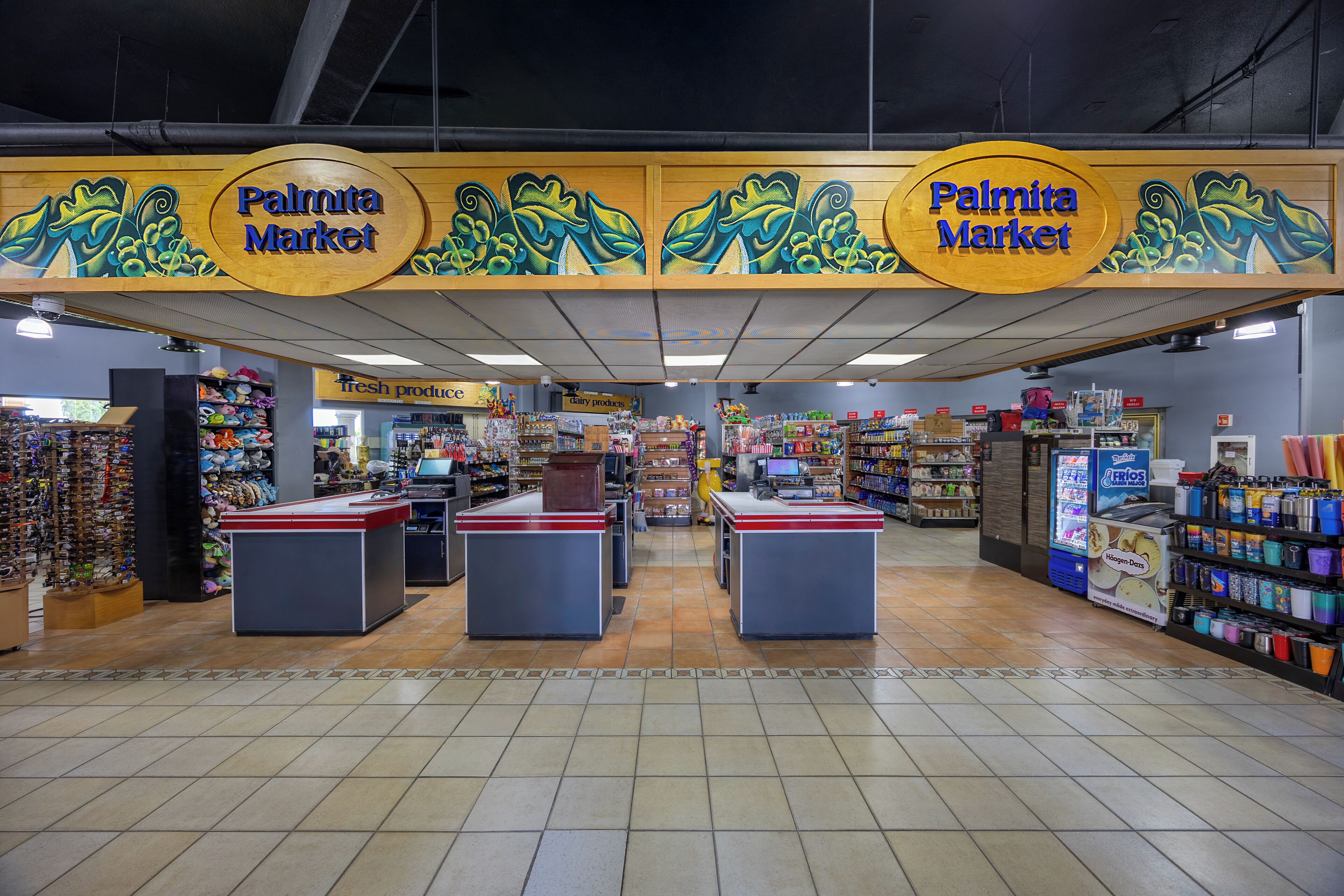 Interior de una tienda de comestibles con dos cajas registradoras, estantes llenos de diversos productos y una sección visible de frutas y verduras frescas.