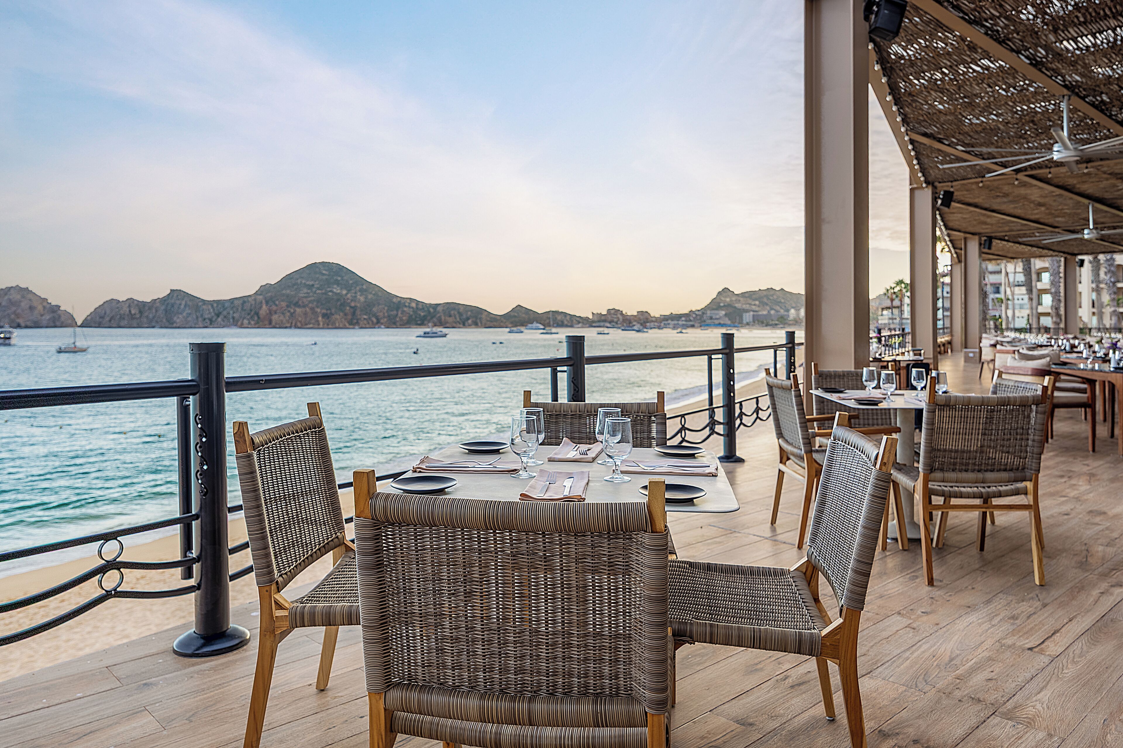 An outdoor dining area with wicker chairs and tables, overlooking a body of water with boats and distant hills.