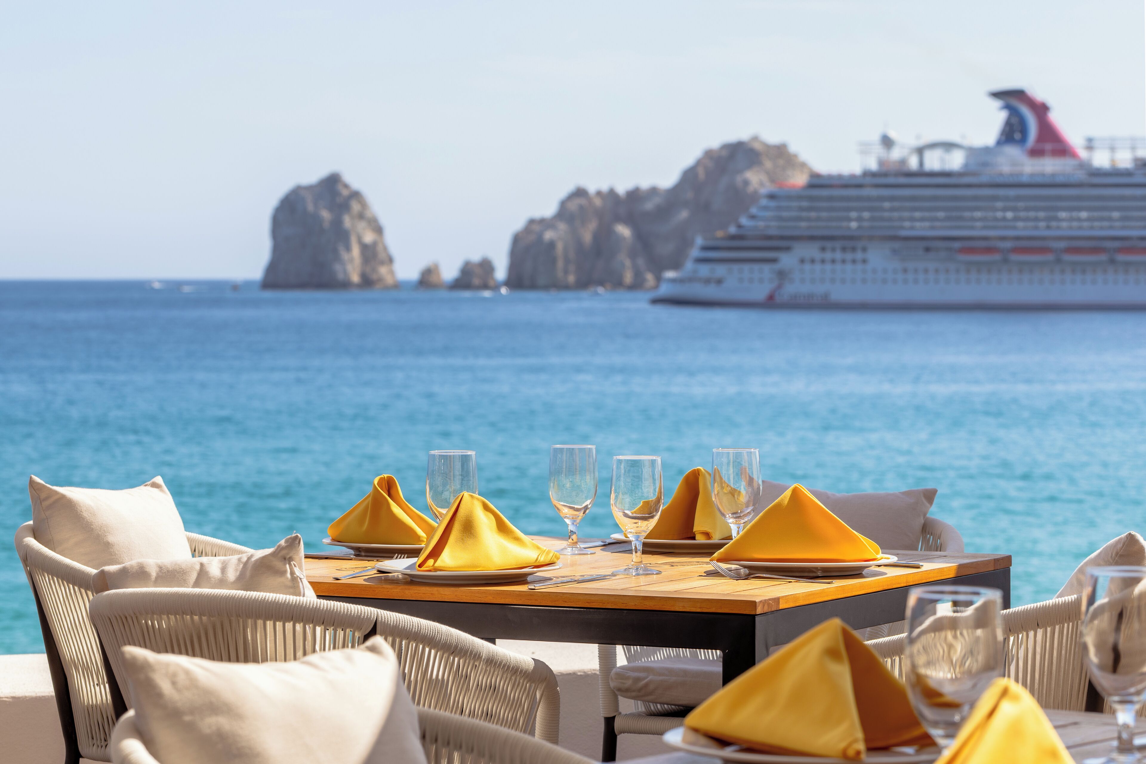 Ein Esszimmertisch ist mit gelben Servietten, Gläsern und Tellern gedeckt, mit Blick aufs Meer und einem Kreuzfahrtschiff im Hintergrund.