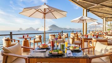 A dining area with tables and chairs, umbrellas, and a view of the sea.