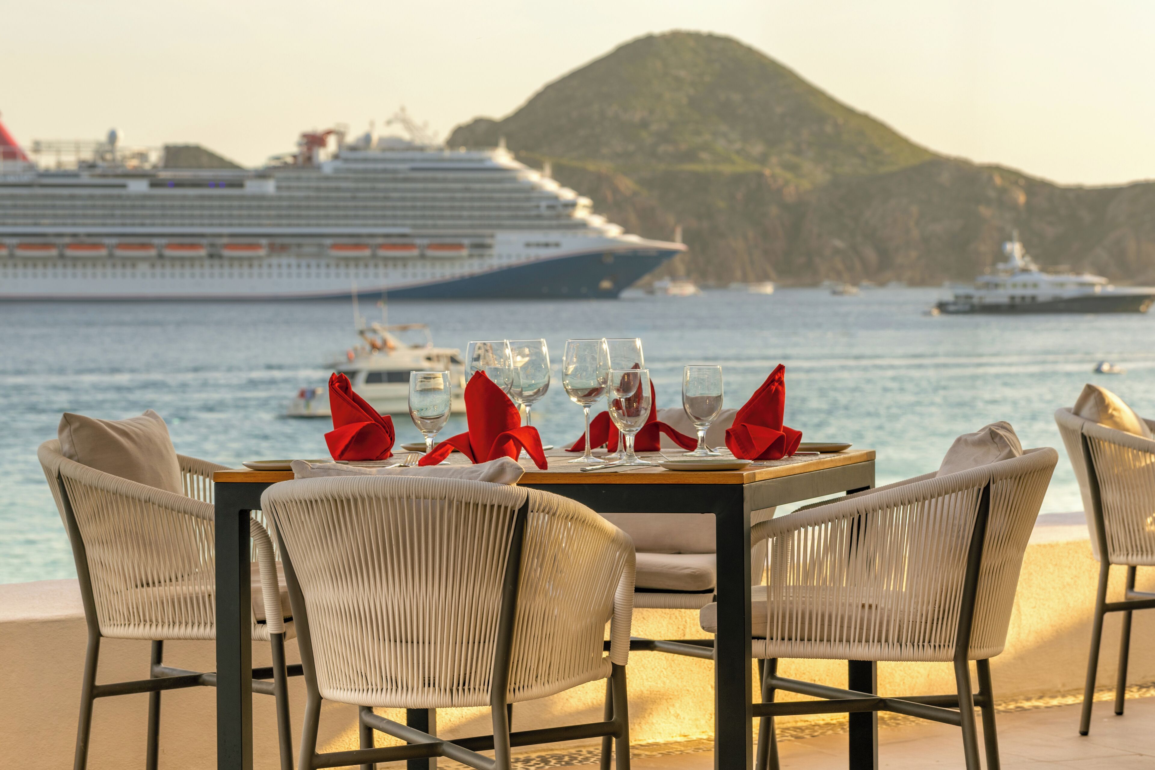 A dining table set with chairs, glasses, and red napkins, overlooking a body of water with cruise ships in the background.