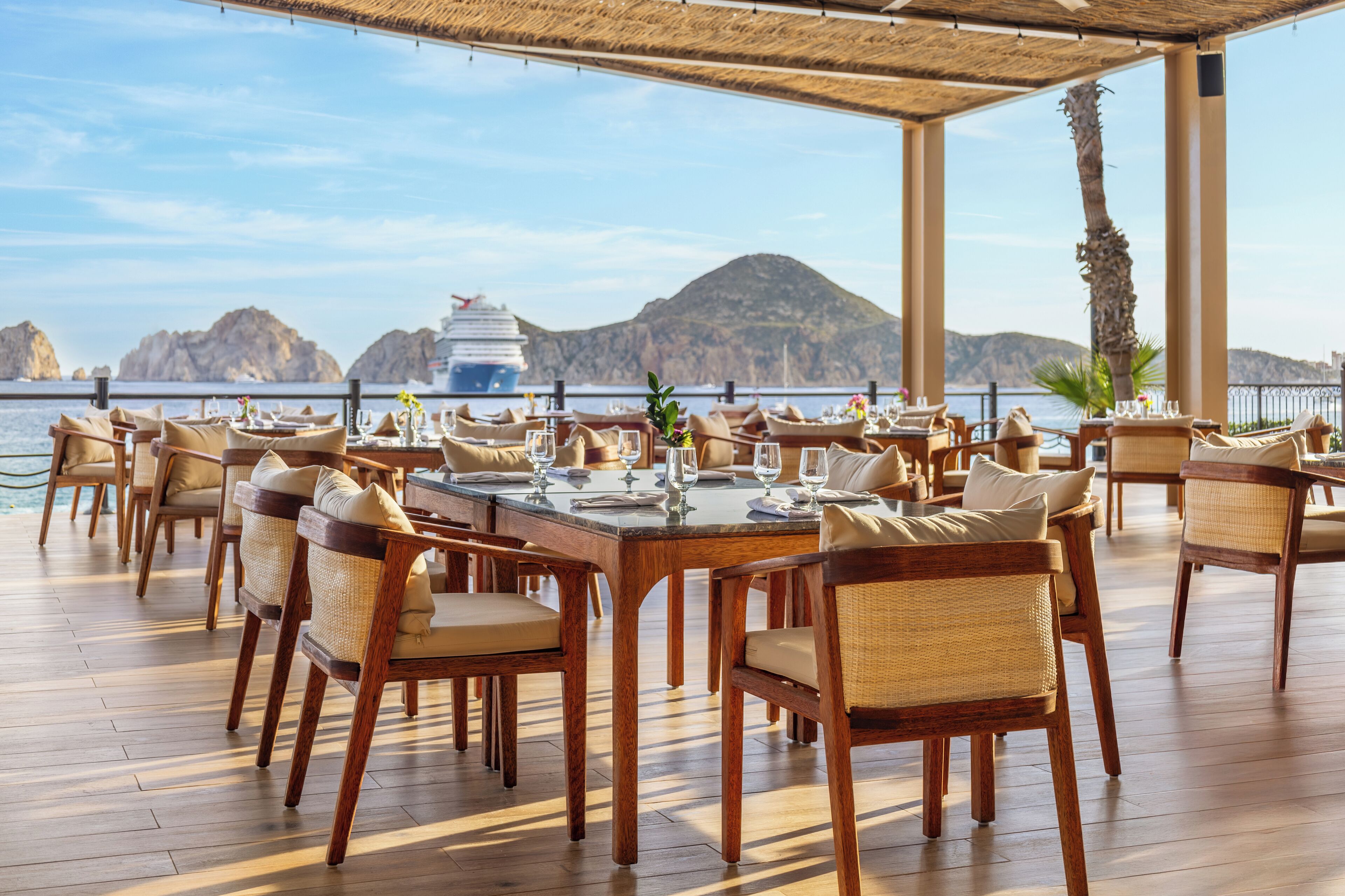 A dining area with wooden tables and chairs, overlooking a body of water with a ship in the background.