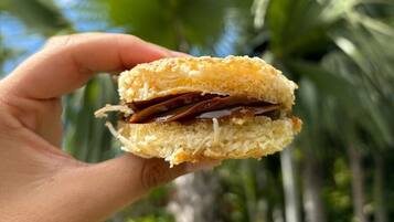 A hand holding a sandwich with a visible filling, palm trees, and greenery in the background.